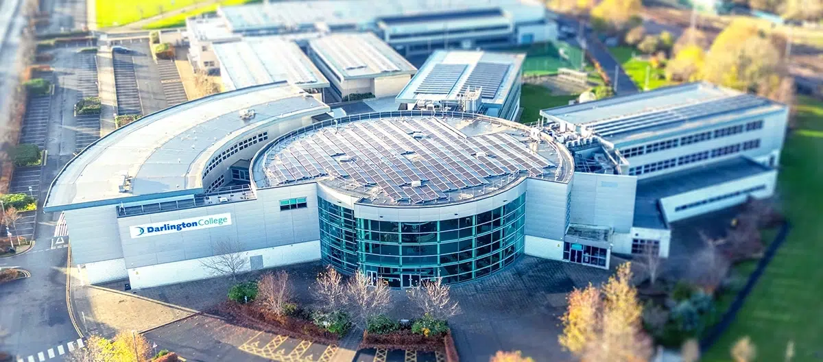 This aerial drone photograph showcases the modern campus of Darlington College. The image features a prominent, semi-circular central building with a large glass facade and a flat roof heavily equipped with solar panels.